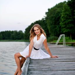 Natalia Mencfeld - Młoda kobieta w białej sukience, siedząca boso na drewnianym molo nad jeziorem, z uśmiechem spogląda w obiektyw, w tle zielone drzewa i błękitne niebo.