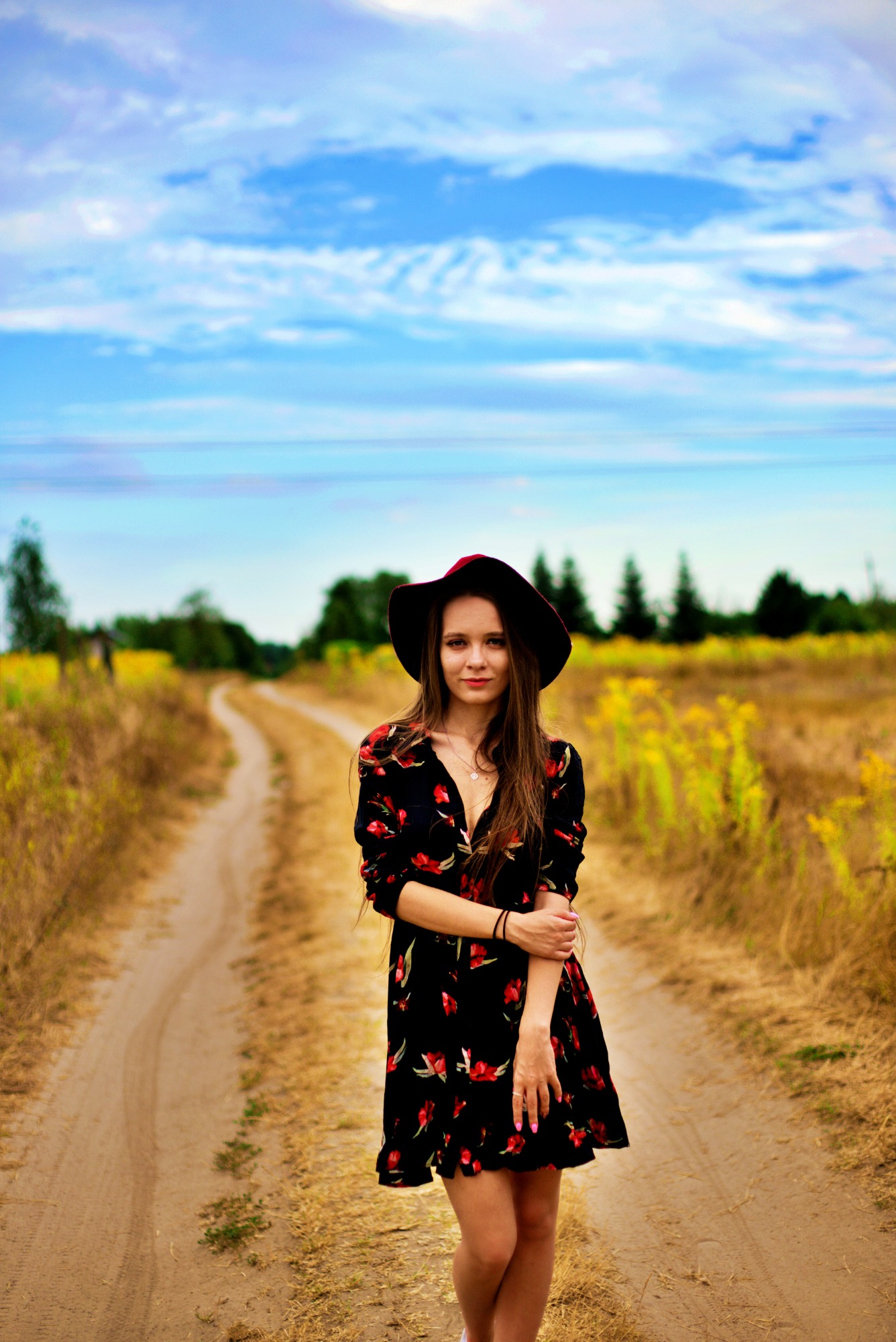 Portret kobiety w czarnej sukience w czerwone kwiaty i kapeluszu na polnej drodze, z niebem w tle.