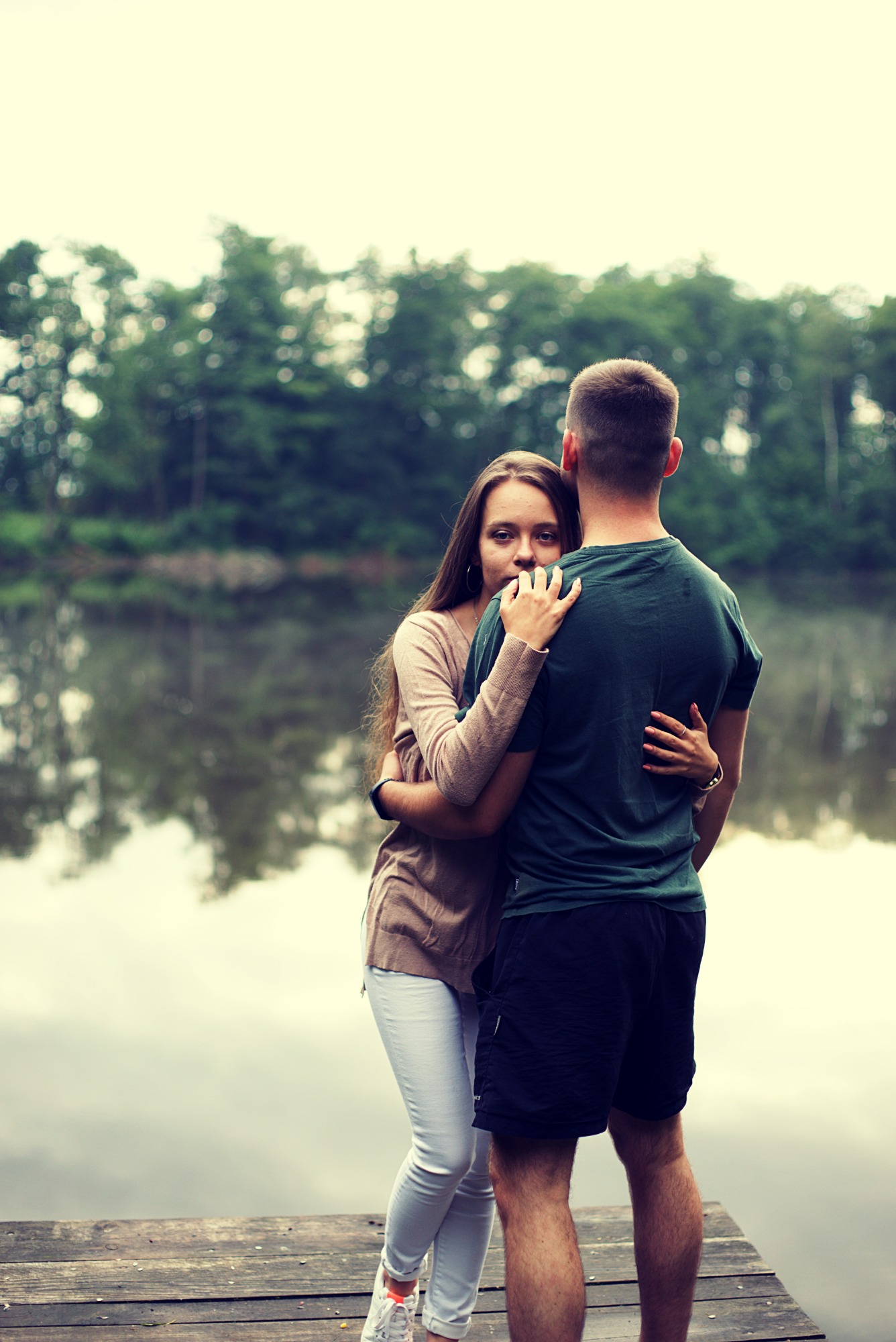 Para obejmująca się na drewnianym pomoście nad jeziorem, kobieta spogląda w obiektyw, w tle widoczny las i odbicie w wodzie.