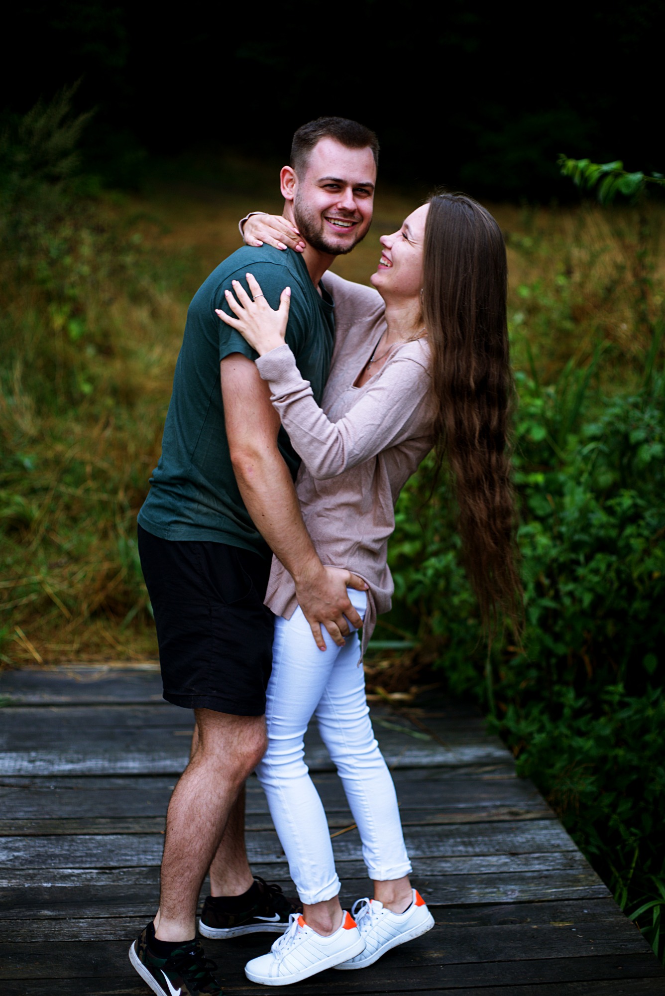 Uśmiechnięta para, mężczyzna w zielonej koszulce i kobieta w beżowym swetrze i białych spodniach, obejmująca się na drewnianym pomoście w plenerze w okolicach Stargardu.