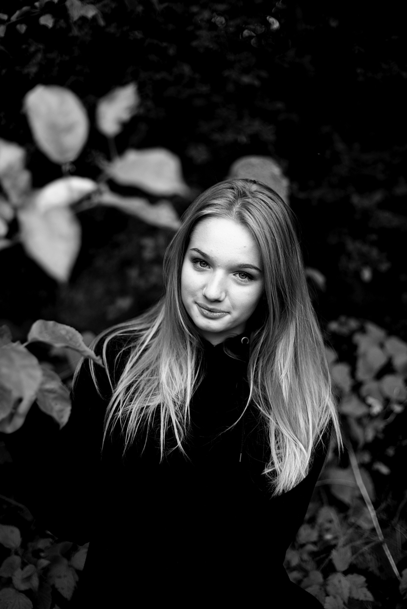 Portret młodej kobiety z długimi włosami, uśmiechającej się w czarno-białym ujęciu na tle liści.
