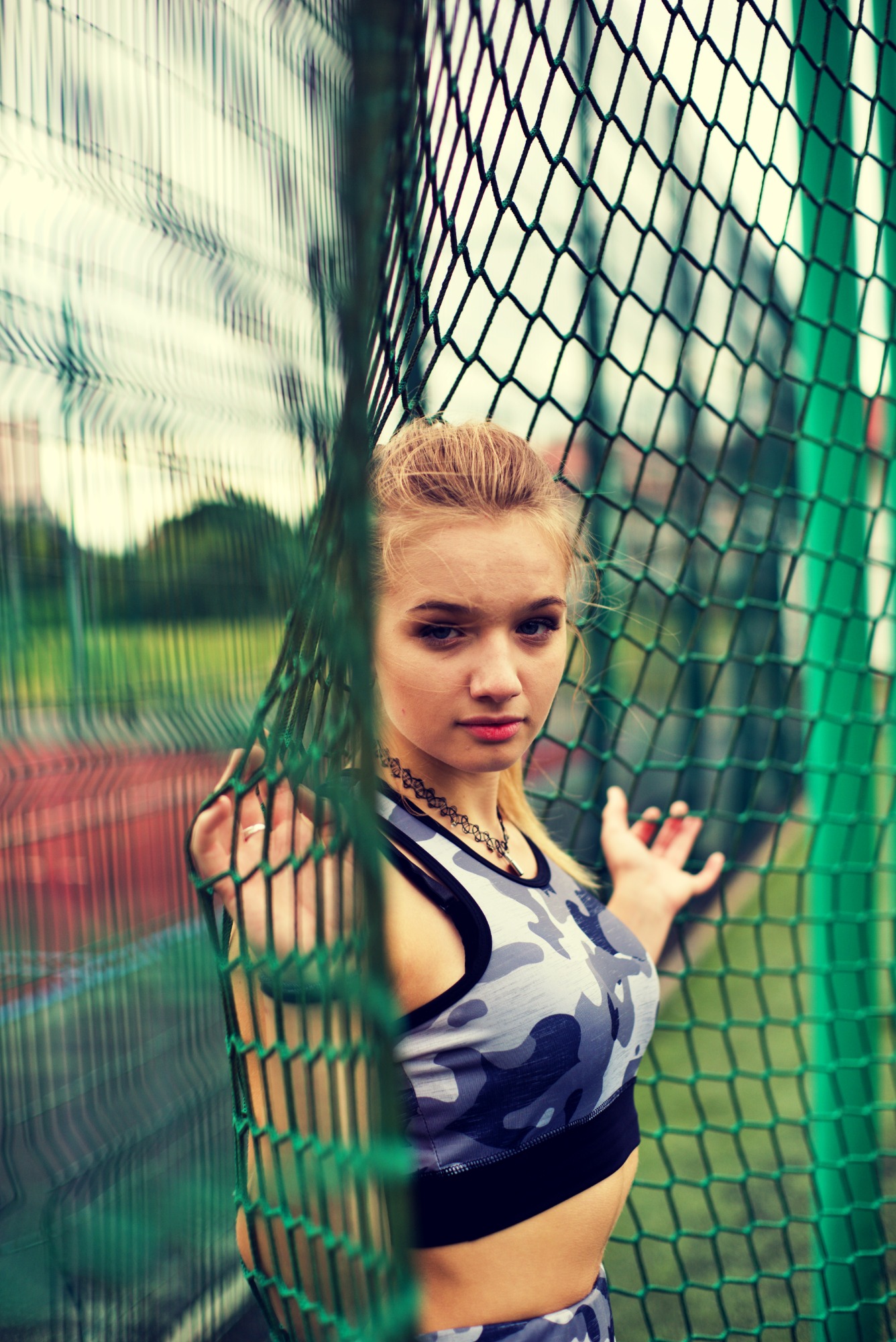 Młoda kobieta w sportowym stroju moro pozuje za zieloną siatką na tle bieżni lekkoatletycznej.