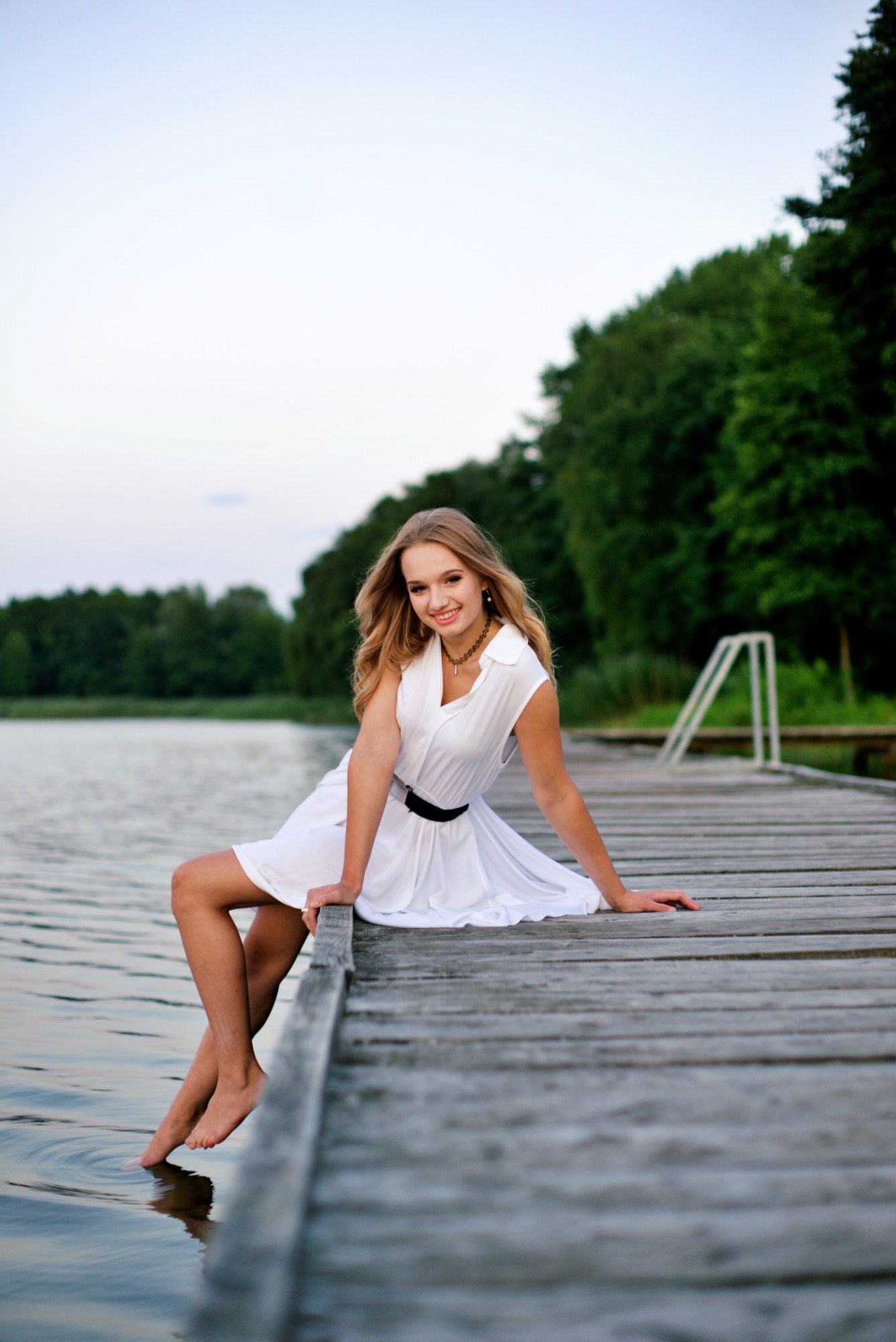 Młoda kobieta w białej sukience, siedząca boso na drewnianym molo nad jeziorem, z uśmiechem spogląda w obiektyw, w tle zielone drzewa i błękitne niebo.