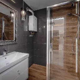 Nowoczesna łazienka z szarymi płytkami, drewnopodobną podłogą i kabiną prysznicową, widoczny bojler i umywalka z szafką, lustro w ozdobnej ramie i dekoracyjna lampa.