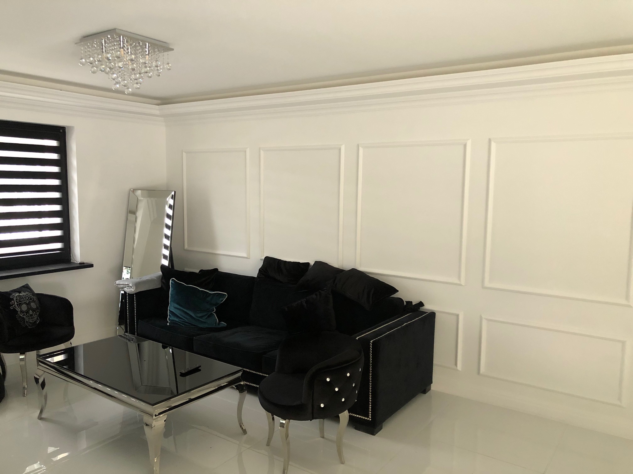 Salon po odnowieniu: białe ściany z dekoracyjnymi listwami, czarna sofa z poduszkami, szklany stolik kawowy na srebrnych nogach i kryształowy żyrandol.