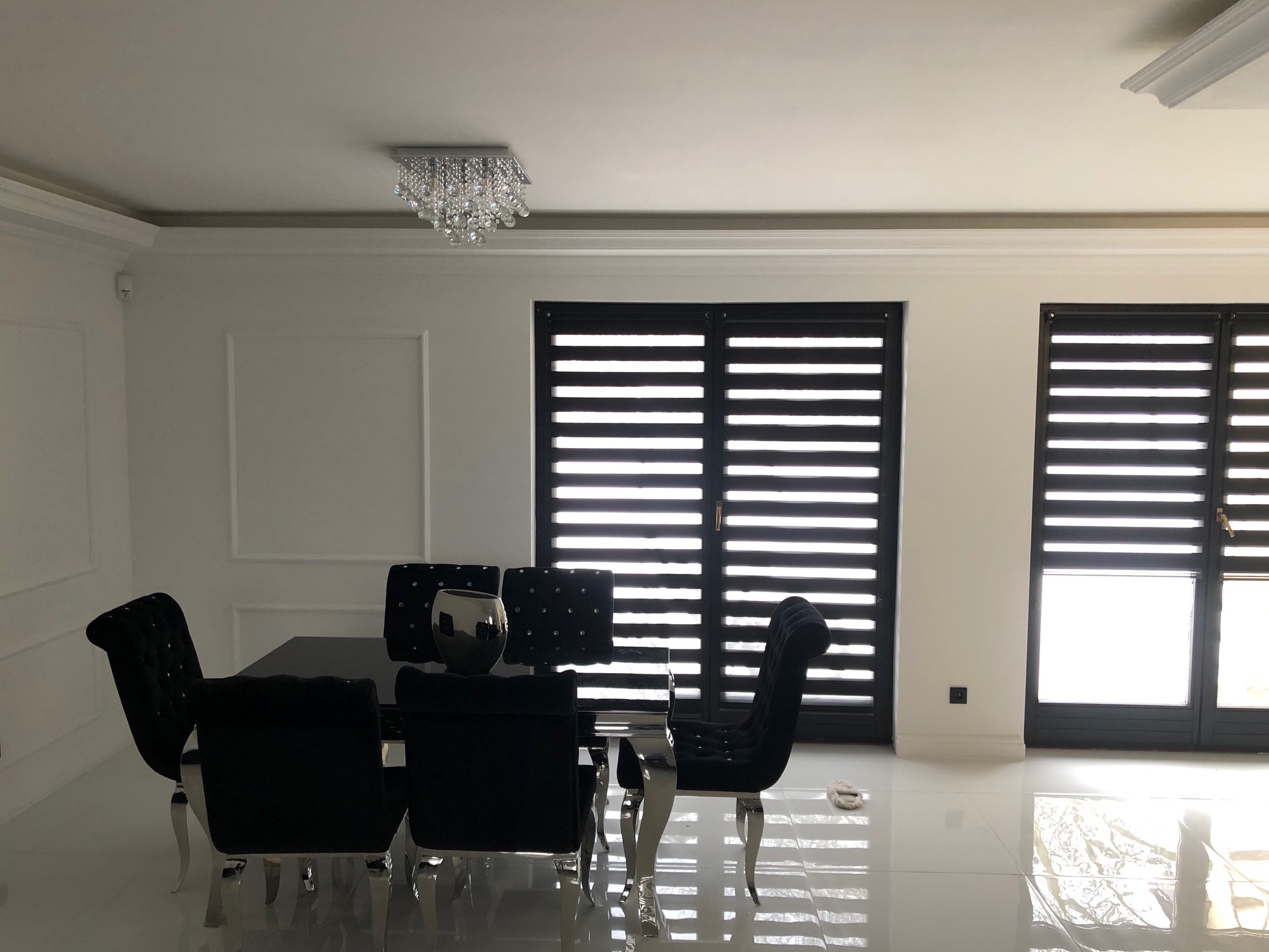 Elegancka jadalnia w bieli i czerni: kryształowy żyrandol, czarne żaluzje okienne, błyszcząca podłoga i zestaw mebli z czarnymi, pikowanymi krzesłami.