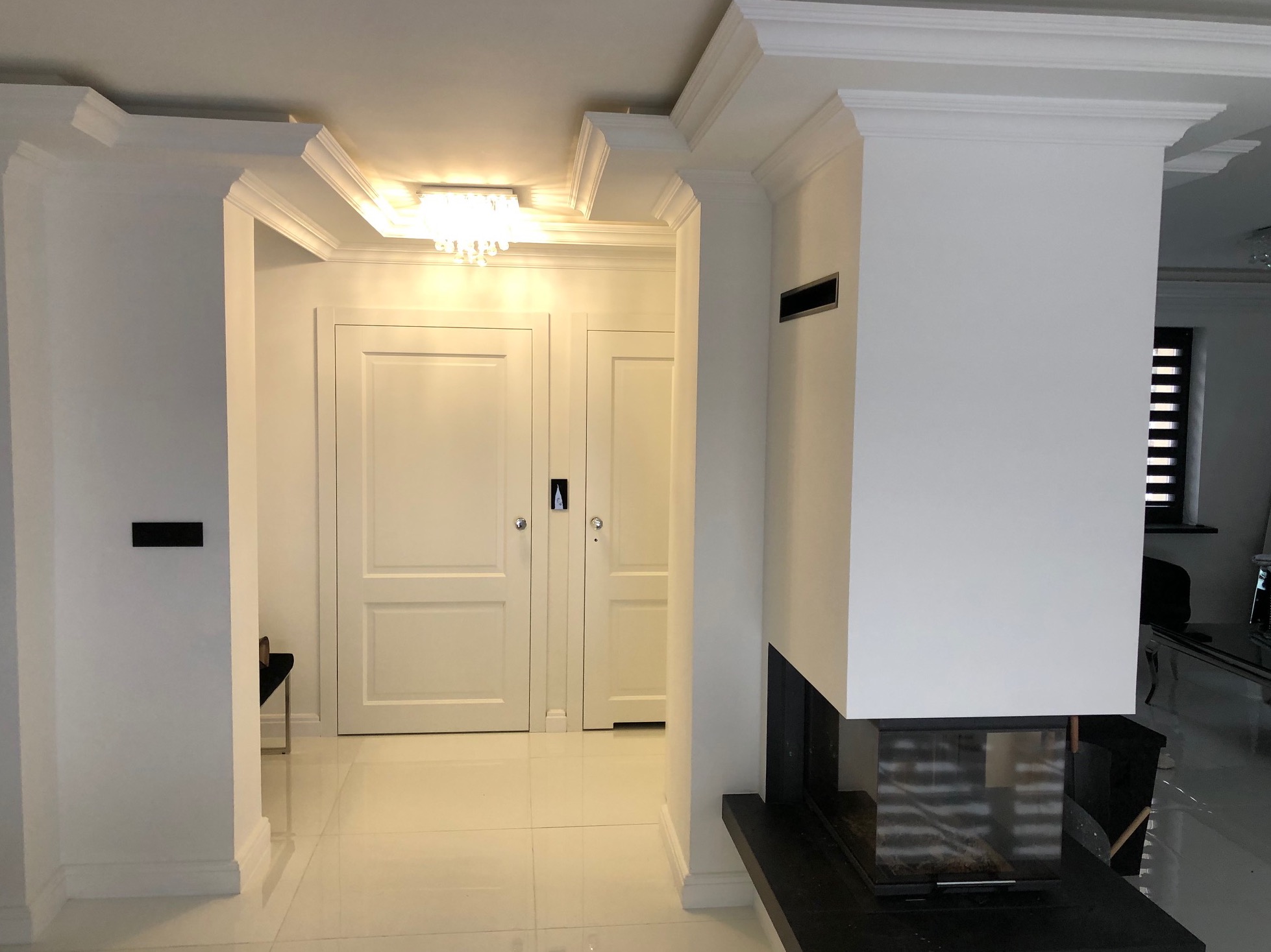 Jasne wnętrze po remoncie: białe ściany z dekoracyjnymi listwami, lśniąca podłoga, drzwi w stylu klasycznym i nowoczesny kominek z czarnym obramowaniem.