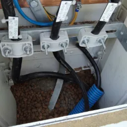 Wnętrze skrzynki elektrycznej z zamocowanymi przewodami oznaczonymi etykietami ZKP TD-ZK YAKXS 4x35 i ZK1-Enelion YKXS 5x10, zabezpieczonej keramzytem i niebieską rurą karbowaną.