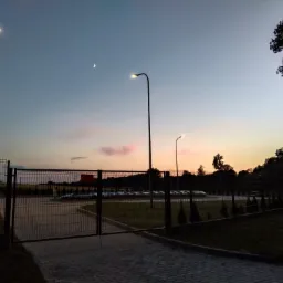 Oświetlona droga dojazdowa z bramą i lampami ulicznymi w tle zachodzącego słońca i księżyca.