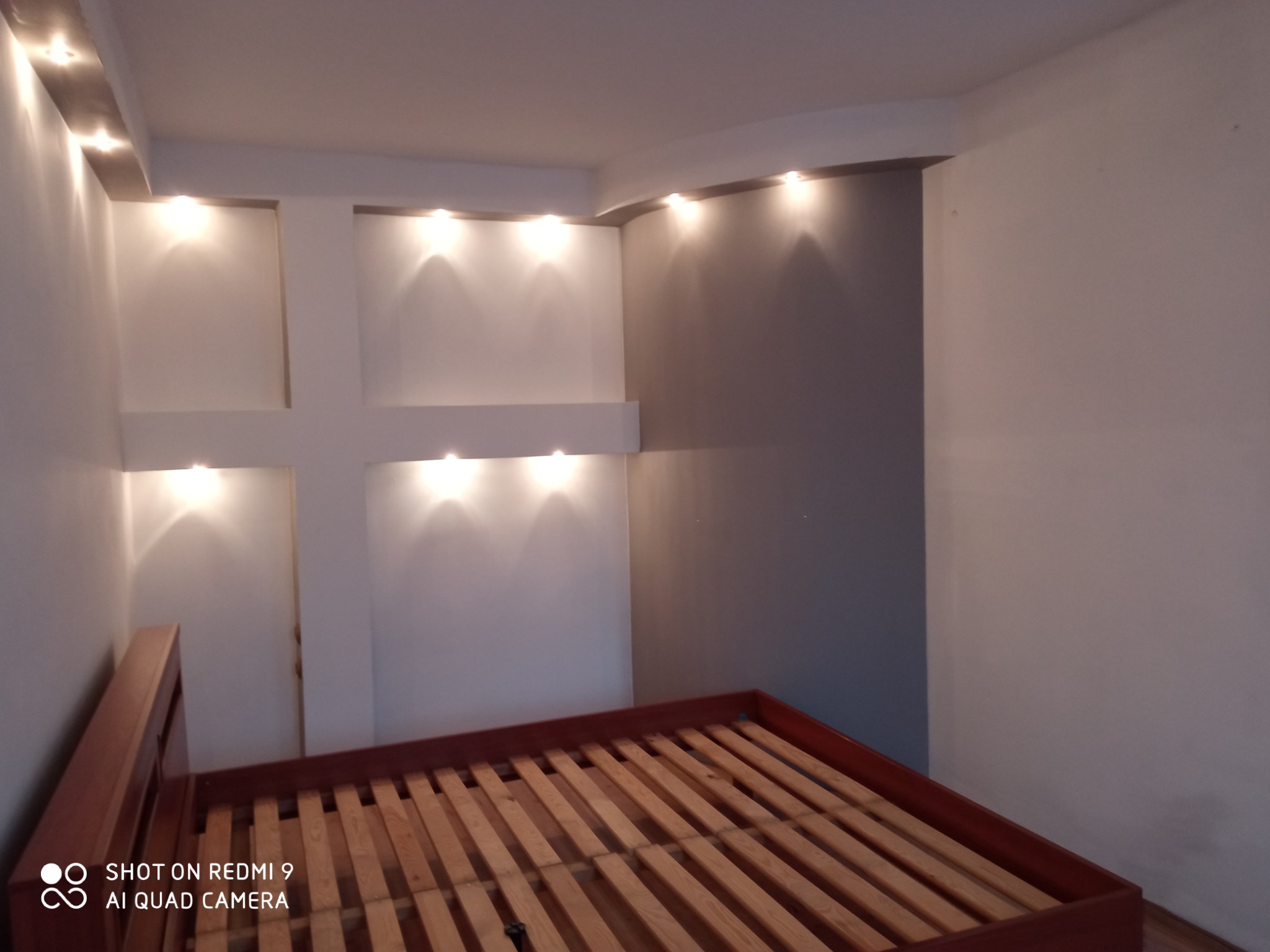 Pokój z zamontowanym oświetleniem punktowym w suficie i ścianie, tworzącym krzyżowy wzór, widoczny szkielet drewnianego łóżka.