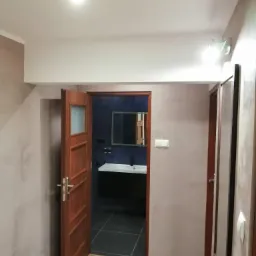 Widok na wykończoną łazienkę z umywalką nablatową i ciemnymi płytkami, widoczny z korytarza z drewnianą podłogą i ścianami w odcieniach beżu i szarości.