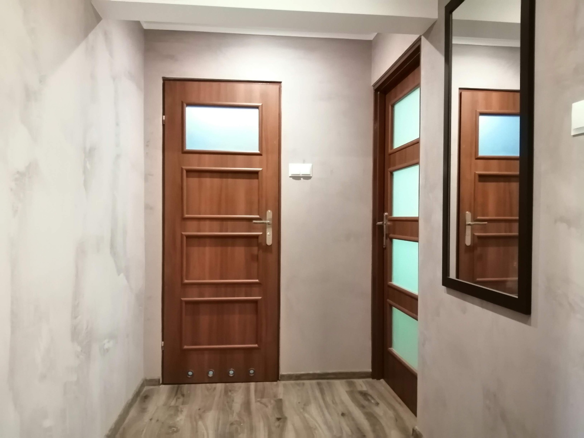 Korytarz po remoncie z dwiema drewnianymi drzwiami z szybami, lustrem w czarnej ramie, ścianami pomalowanymi z efektem chmur i podłogą imitującą drewno.