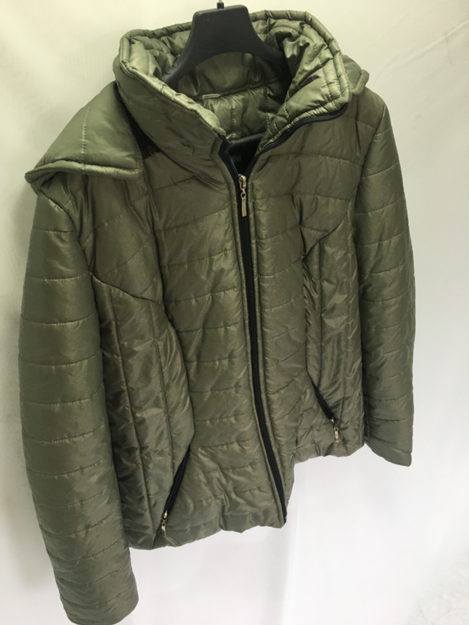 Oliwkowa, pikowana kurtka damska z kapturem, wisząca na czarnym wieszaku na tle białej ściany, z widocznym zamkiem błyskawicznym i kieszeniami.