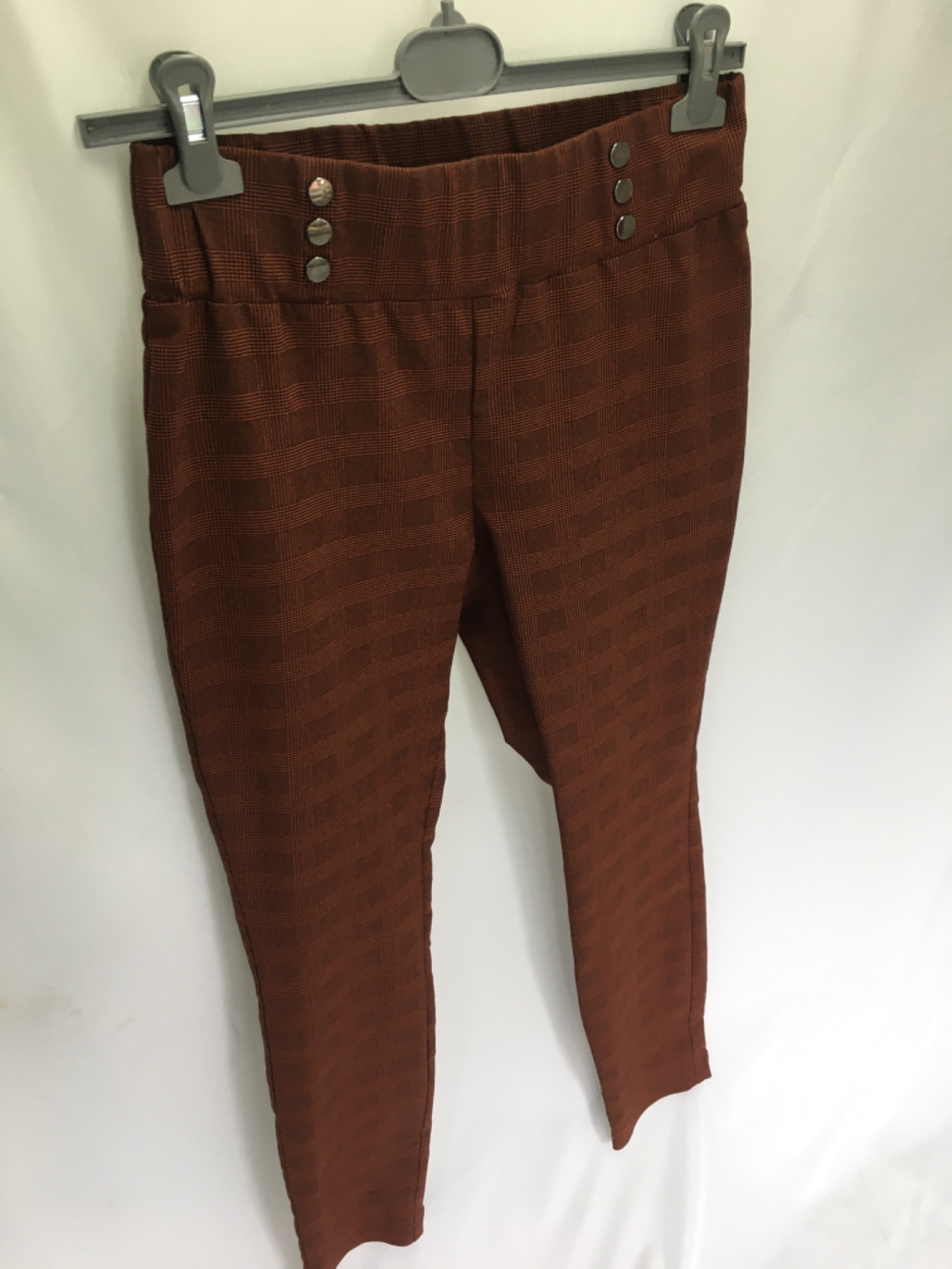 Brązowe spodnie damskie w drobną kratę, z wysokim stanem zdobionym trzema guzikami po każdej stronie, wiszące na wieszaku na białym tle.