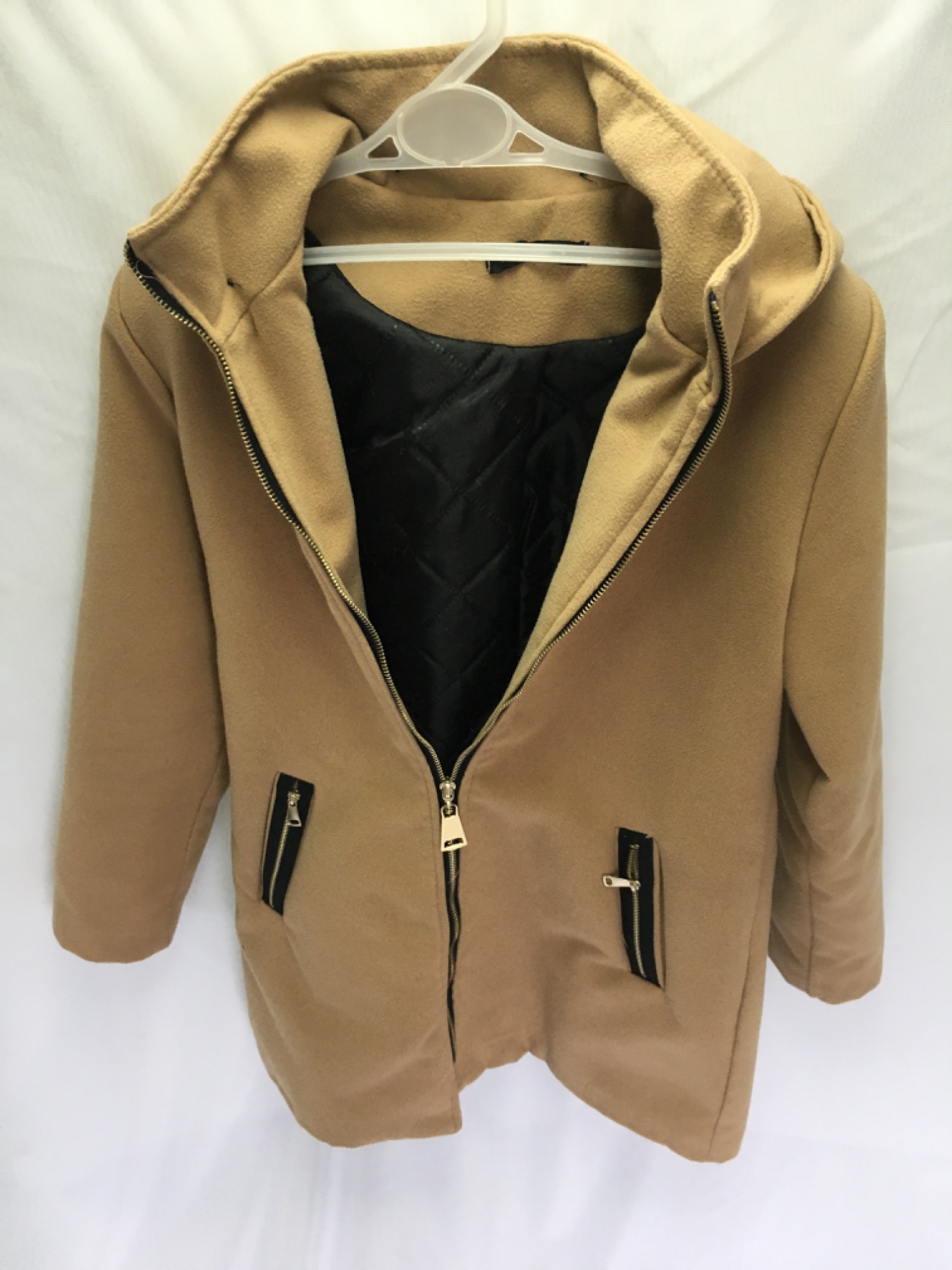 Beżowy, rozpięty płaszcz damski z czarną, pikowaną podszewką, wiszący na plastikowym wieszaku na tle białej tkaniny.