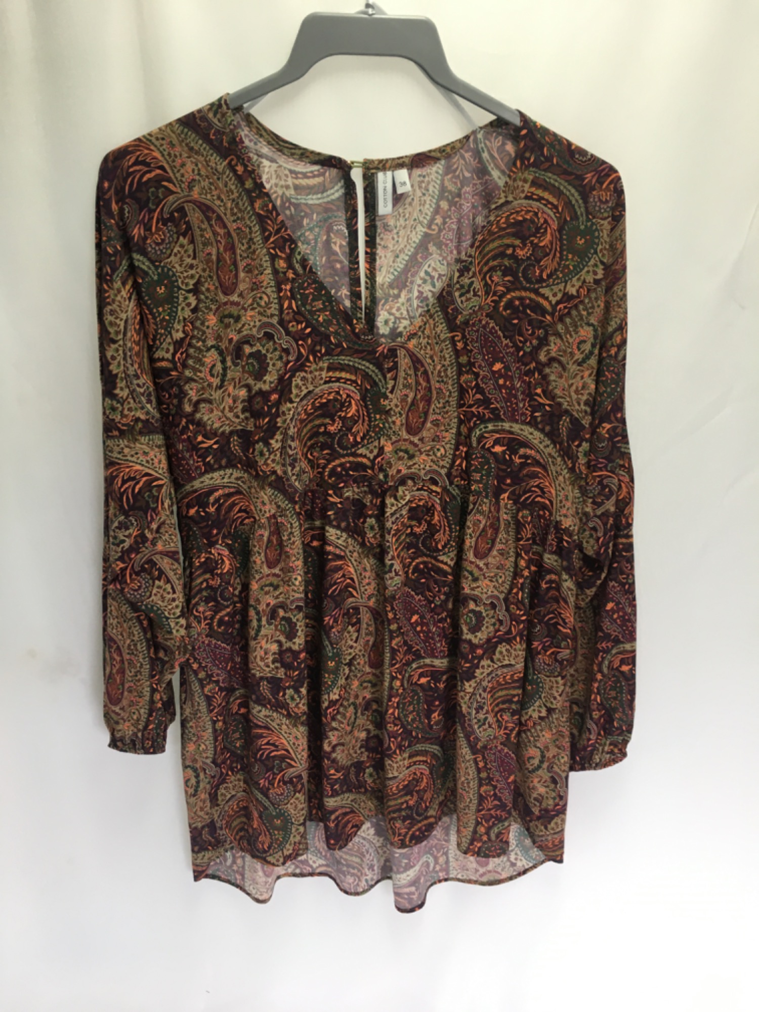 Bluzka damska z długim rękawem w etniczny wzór paisley w odcieniach brązu, pomarańczy i zieleni, wisząca na szarym wieszaku na tle białej ściany.