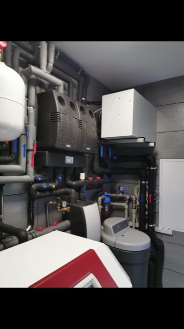 Kompletna instalacja grzewcza w kotłowni z dwoma czarnymi zbiornikami akumulacyjnymi, białym zasobnikiem, szarym zmiękczaczem wody i licznymi rurami izolowanymi szarą pianką.