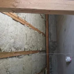 Ocieplenie poddasza wełną mineralną, widoczne drewniane belki konstrukcyjne i sznurek do podtrzymywania izolacji, na podłodze leży rolka sznurka.