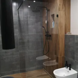 Nowoczesna łazienka z kabiną prysznicową oddzieloną szklaną ścianką, czarną armaturą i płytkami imitującymi drewno na jednej ze ścian.