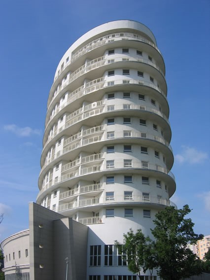 Nowoczesny, biały budynek mieszkalny z zaokrąglonymi balkonami w Krakowie, widoczny na tle błękitnego nieba.