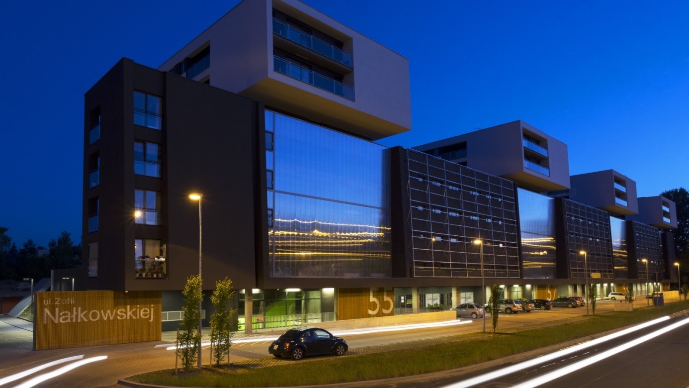 Nowoczesny budynek apartamentowy z dużymi przeszkleniami i balkonami, oświetlony nocą, z widocznymi światłami samochodów i zielenią.