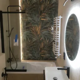 Nowoczesna łazienka z wanną, czarną armaturą, umywalką nablatową i podświetlanym lustrem owalnym, płytki z motywem tropikalnych liści na ścianie.