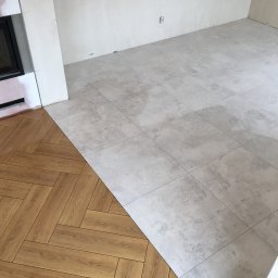 ZAKŁAD REMONTOWO-BUDOWLANY RYSZARD KORBUT - Fragment podłogi w trakcie łączenia dwóch rodzajów wykończenia: jasnoszare płytki kwadratowe i panele drewnopodobne ułożone w jodełkę, widoczne ślady po fugowaniu na płytkach, surowa ściana w tle.
