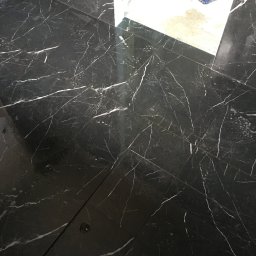 ZAKŁAD REMONTOWO-BUDOWLANY RYSZARD KORBUT - Błyszcząca, czarna podłoga z płytek imitujących marmur z białymi żyłkami, świeżo po umyciu, z widocznymi smugami wody i kroplami na powierzchni.