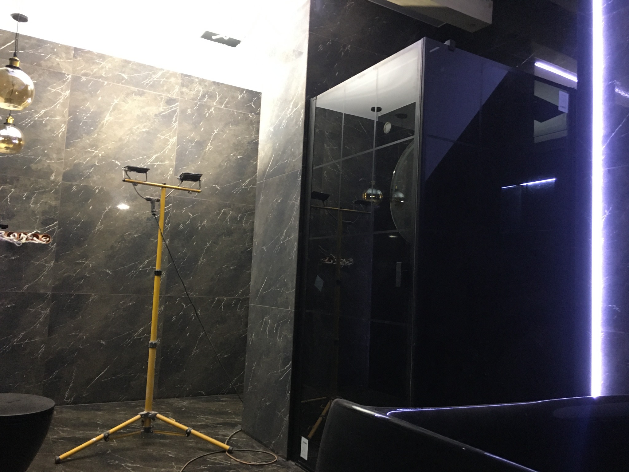 Ciemna łazienka w Świdnicy z marmurowymi płytkami, oświetlona halogenem na statywie, z widoczną kabiną prysznicową i fragmentem wanny.