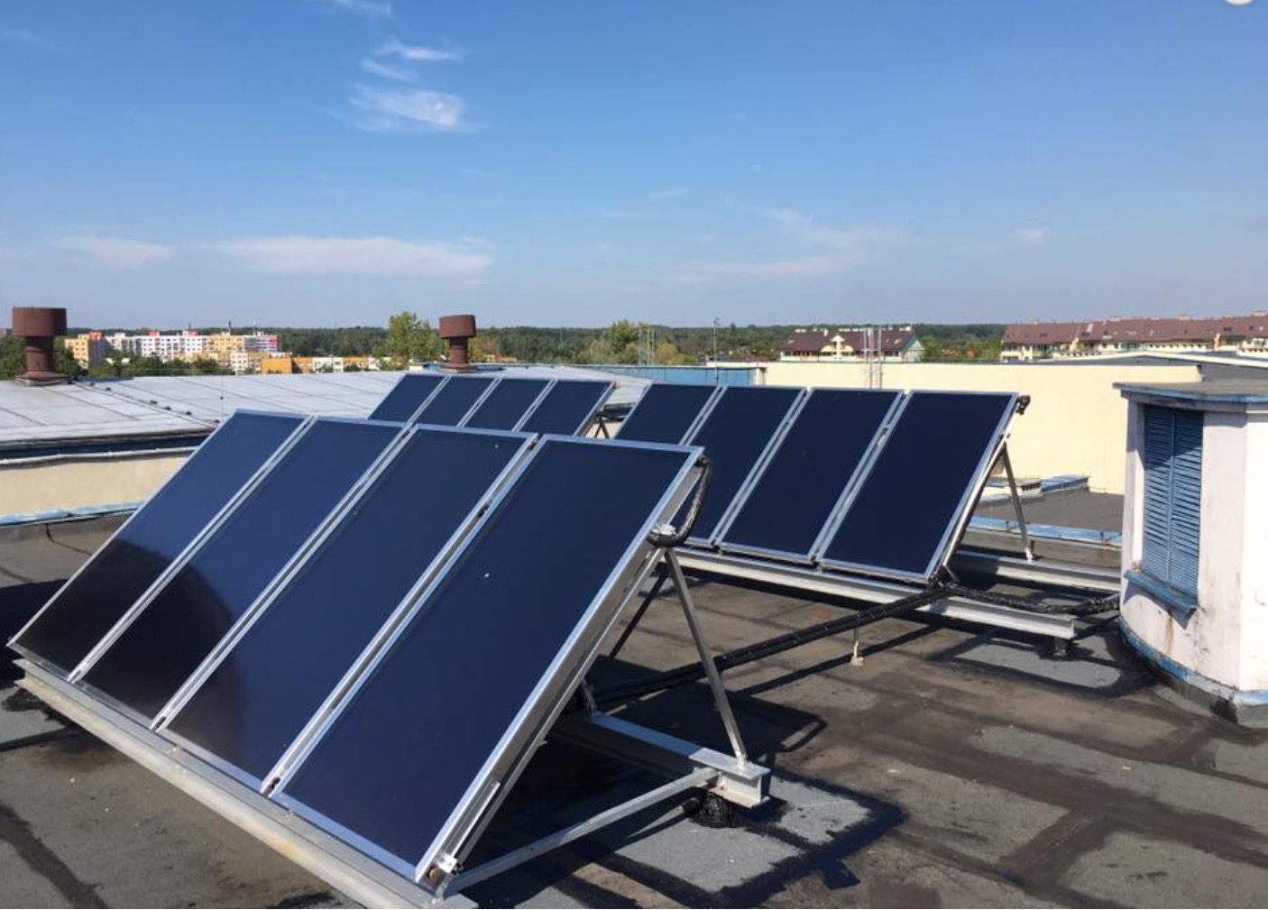 Panele słoneczne na płaskim dachu budynku z widokiem na miasto w tle, błękitne niebo.
