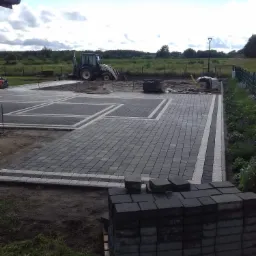 Układanie kostki brukowej w geometryczny wzór z jasnymi obwódkami, widoczny traktor i ubijarka w tle, paleta z kostką na pierwszym planie.