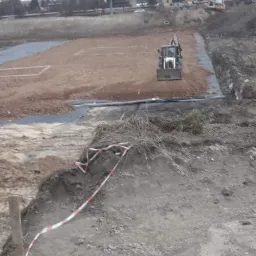 Teren budowy z widocznym traktorem wyrównującym grunt pod przyszłą inwestycję, zabezpieczony taśmą ostrzegawczą.