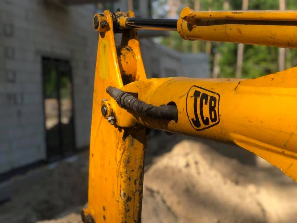 Żółte ramię koparki JCB z widocznymi śladami użytkowania i logo, na tle ściany budynku w budowie i drzew