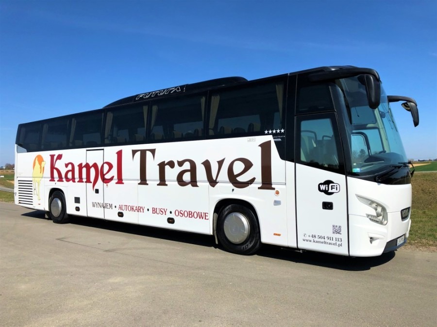 Biały autokar turystyczny marki Futura z brązowym napisem 'Kamel Travel' i sylwetką wielbłąda, zaparkowany na asfalcie w słoneczny dzień, z widocznym logo Wi-Fi i danymi kontaktowymi firmy.