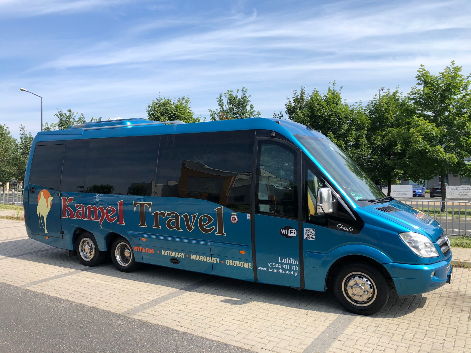Niebieski minibus firmy Kamel Travel z logo z wielbłądem, zaparkowany na placu z kostki brukowej w słoneczny dzień.