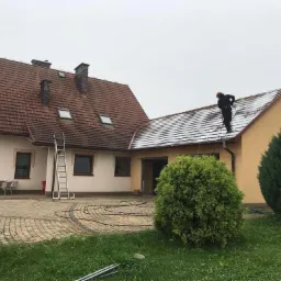 Pracownik w kombinezonie i kasku, stojący na dachu domu jednorodzinnego i czyszczący go myjką ciśnieniową. Część dachu jest już oczyszczona, widoczny kontrast między czystą a zabrudzoną...