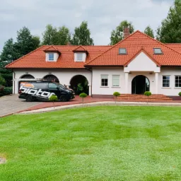 Dwupiętrowy dom z czerwonym dachem, na podjeździe stoi czarny samochód dostawczy z logo firmy 'Biotor' i napisem 'Malowanie Dachów', na dachu widoczna drabina.