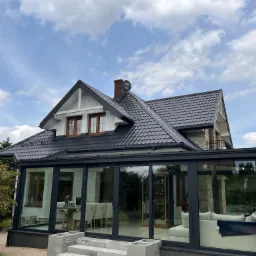 Dom jednorodzinny z ciemnoszarą dachówką ceramiczną i przeszkloną oranżerią, widok z zewnątrz w słoneczny dzień. Na dachu widoczna antena satelitarna. W tle błękitne niebo z chmurami.