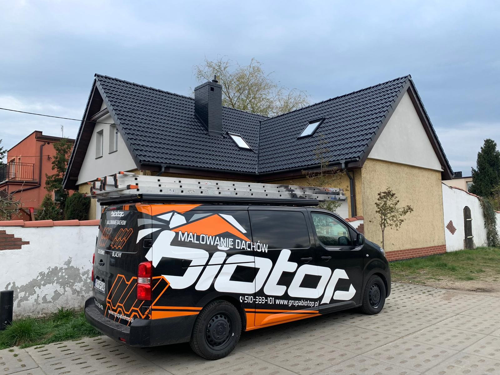 Czarny van z logo firmy dekarskiej i hasłem 'Malowanie Dachów' zaparkowany przed domem z nowo pomalowanym, ciemnym dachem ceramicznym i kominem. Na dachu samochodu drabina.