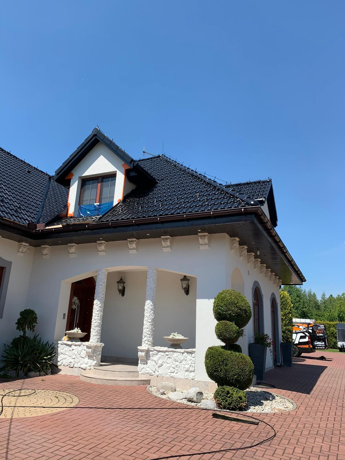 Dom jednorodzinny z nowym, czarnym dachem z dachówki ceramicznej. Na dachu widoczne zabezpieczenie okna połaciowego folią. Na podjeździe kostka brukowa.
