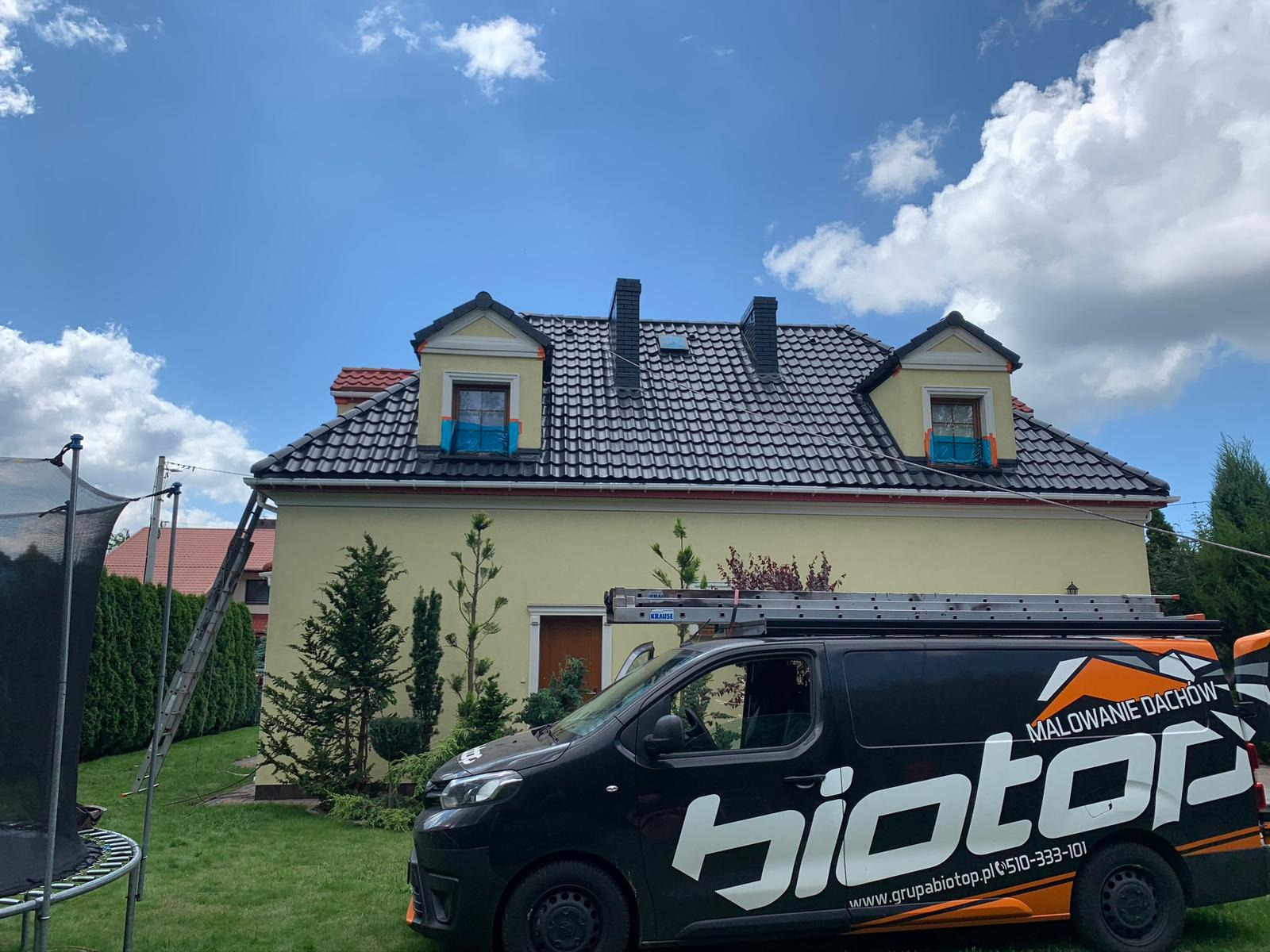 Świeżo pomalowany dach z czarną dachówką na żółtym domu z zabezpieczonymi oknami, drabina oparta o rynnę, firmowy van z napisem 'Malowanie Dachów biotop' na trawniku.