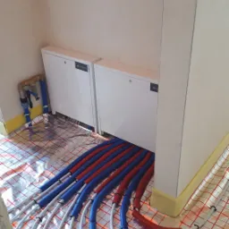 Rozłożone rury ogrzewania podłogowego w kolorze czerwonym i niebieskim na folii aluminiowej z siatką, przed szafkami rozdzielczymi z zaworami.