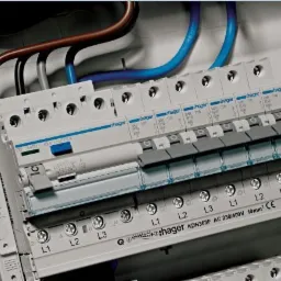 Szczegółowe ujęcie tablicy rozdzielczej Hager z widocznymi bezpiecznikami, przewodami w kolorach niebieskim i brązowym, oznaczeniami L1, L2, L3 oraz żółtym symbolem ostrzegawczym.