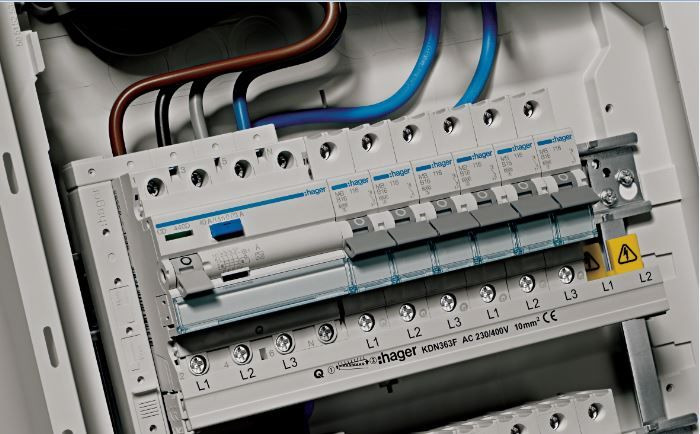 Szczegółowe ujęcie tablicy rozdzielczej Hager z widocznymi bezpiecznikami, przewodami w kolorach niebieskim i brązowym, oznaczeniami L1, L2, L3 oraz żółtym symbolem ostrzegawczym.