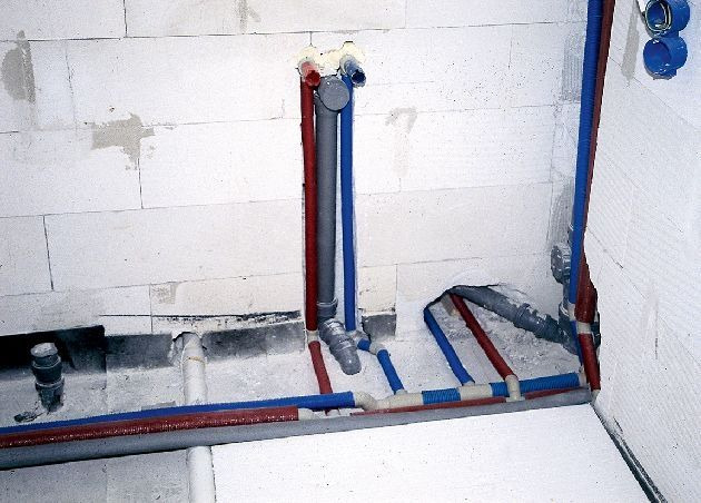 Surowe ściany z betonu komórkowego z widocznymi instalacjami wodno-kanalizacyjnymi: rury w kolorze czerwonym, niebieskim i szarym, prowadzone w podłodze i ścianie, w trakcie montażu.