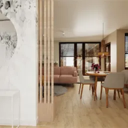 Jasne wnętrze mieszkania w Wilanowie: okrągłe lustro na ścianie z motywem roślinnym, drewniany ażurowy przepierzenie, różowa sofa, okrągły stół z kwiatami i krzesłami, biała zabudowa kuchenna...