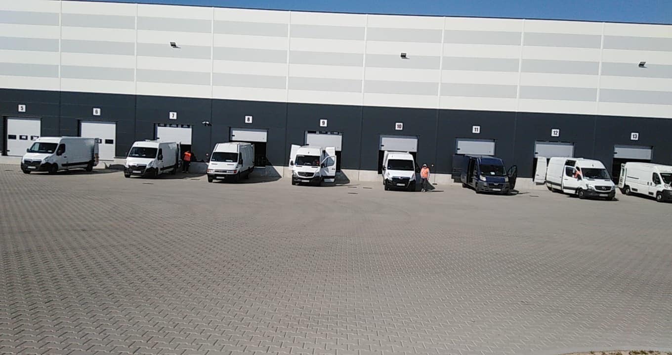 Flota busów dostawczych zaparkowana przed rampami magazynowymi, niektóre z otwartymi drzwiami, sugerująca załadunek lub rozładunek towaru.