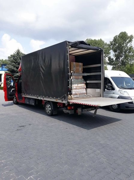 Czerwony samochód dostawczy z otwartą przestrzenią ładunkową, załadowany kartonami i zabezpieczonym towarem, widoczny na parkingu obok białego busa.