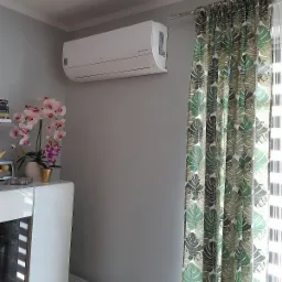 Biały klimatyzator LG Dual Inverter zamontowany na szarej ścianie w pokoju z zasłonami w liście monstery i białym regałem z orchideą.