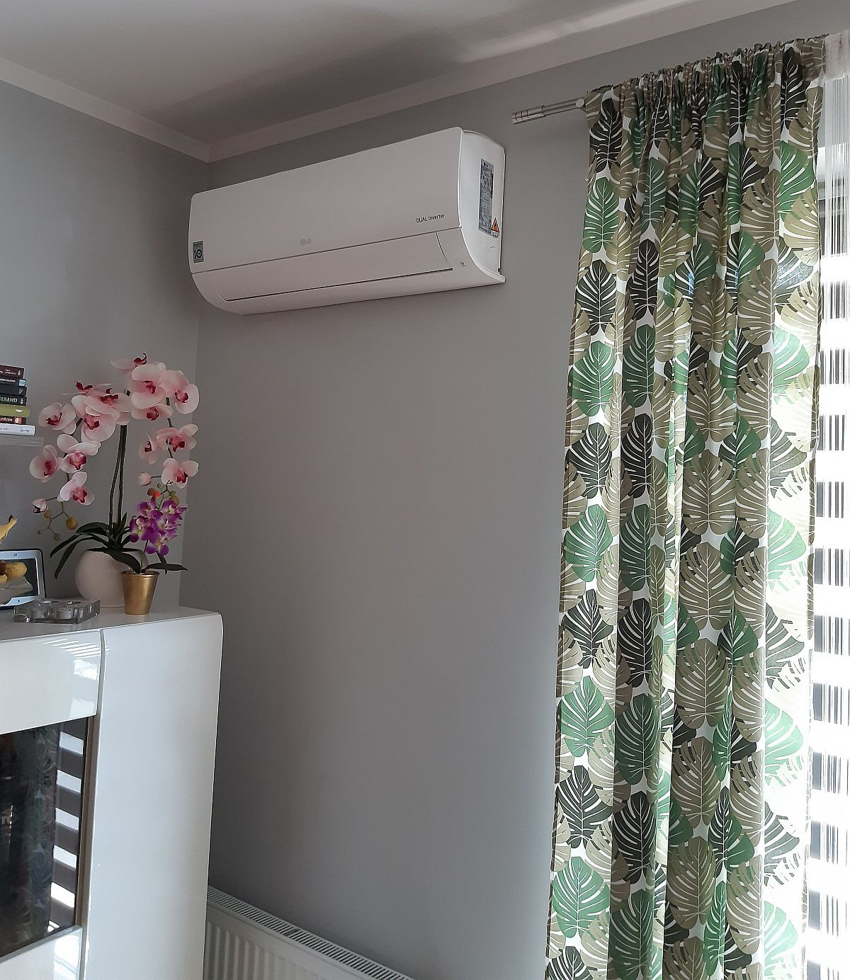 Biały klimatyzator LG Dual Inverter zamontowany na szarej ścianie w pokoju z zasłonami w liście monstery i białym regałem z orchideą.